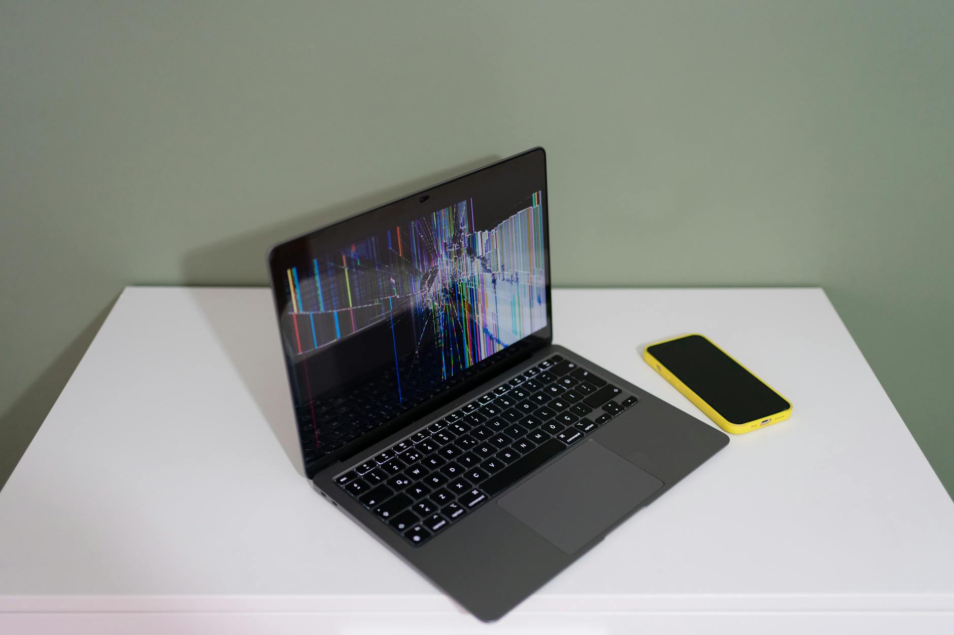 Uszkodzony laptop z kolorowymi liniami obok żółtego smartfona na biurku